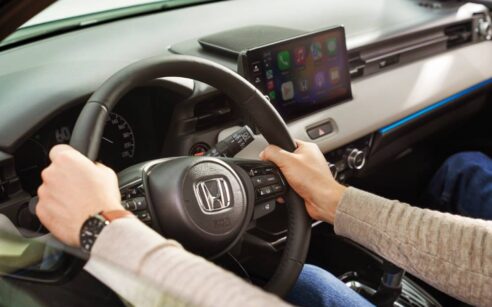 Mãos de um condutor ao volante de um automóvel Honda, com o painel de instrumentos e o ecrã multimédia ao centro