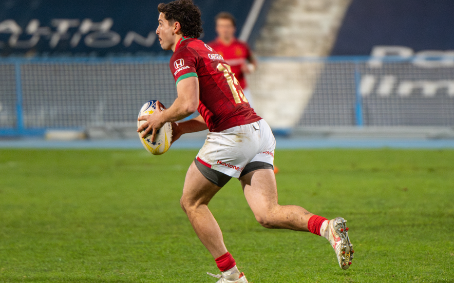 Jogador de râguebi da seleção portuguesa corre com bola em campo, equipamento vermelho, logótipo Honda visível.