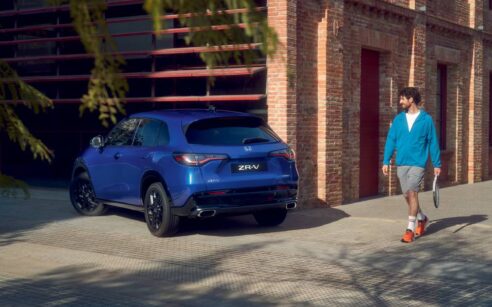 ZR-V azul estacionado junto a um edifício, enquanto um homem caminha no passeio e segura uma raquete, em ambiente urbano e iluminado pela luz natural.