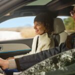 Duas pessoas adultas dentro de um automóvel em movimento; um homem conduz enquanto uma mulher segue no lugar do passageiro, ambos sorridentes, com paisagem rural visível através das janelas.