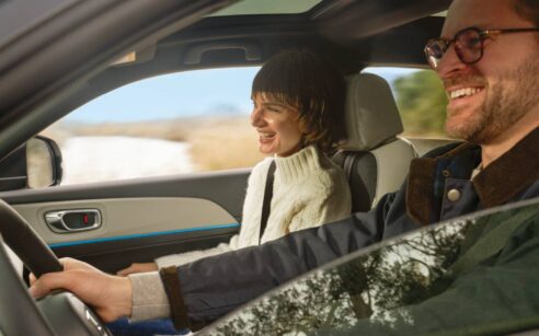 Duas pessoas adultas dentro de um automóvel em movimento; um homem conduz enquanto uma mulher segue no lugar do passageiro, ambos sorridentes, com paisagem rural visível através das janelas.