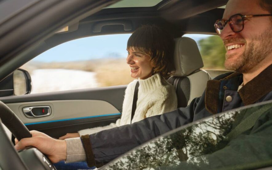 Duas pessoas adultas dentro de um automóvel em movimento; um homem conduz enquanto uma mulher segue no lugar do passageiro, ambos sorridentes, com paisagem rural visível através das janelas.