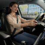 Mãe a conduzir um carro com expressão tranquila, usando cinto de segurança, enquanto uma criança sorri sentada no banco traseiro; ao fundo, vê-se um ambiente urbano através da janela.