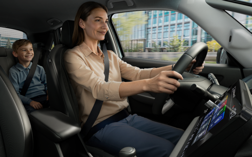 Mãe a conduzir um carro com expressão tranquila, usando cinto de segurança, enquanto uma criança sorri sentada no banco traseiro; ao fundo, vê-se um ambiente urbano através da janela.