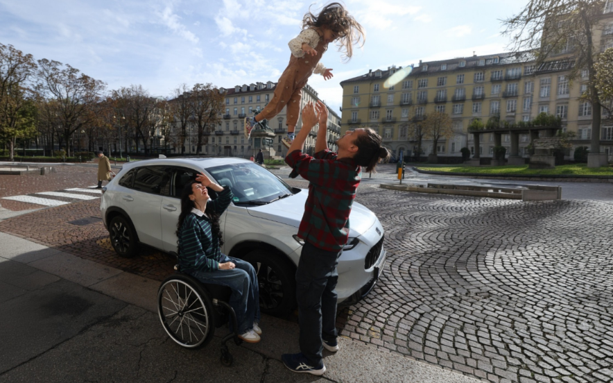 Família junto a um SUV branco numa praça urbana; uma pessoa em cadeira de rodas observa outra a levantar uma criança no ar ao lado de um Honda
