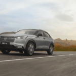 Automóvel SUV cinzento a circular numa estrada ampla, com vegetação nas margens e céu nublado ao pôr do sol.