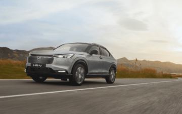 Automóvel SUV cinzento a circular numa estrada ampla, com vegetação nas margens e céu nublado ao pôr do sol.