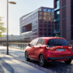 Honda Jazz vermelho a circular numa rua urbana, junto a edifícios modernos, ao pôr do sol, com duas pessoas a caminhar no passeio.
