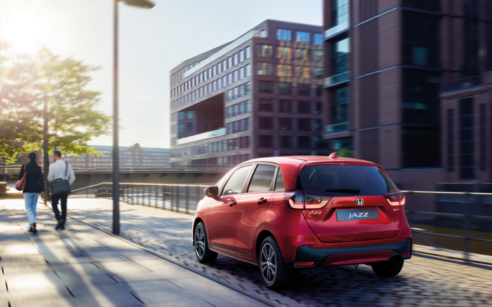 Honda Jazz vermelho a circular numa rua urbana, junto a edifícios modernos, ao pôr do sol, com duas pessoas a caminhar no passeio.